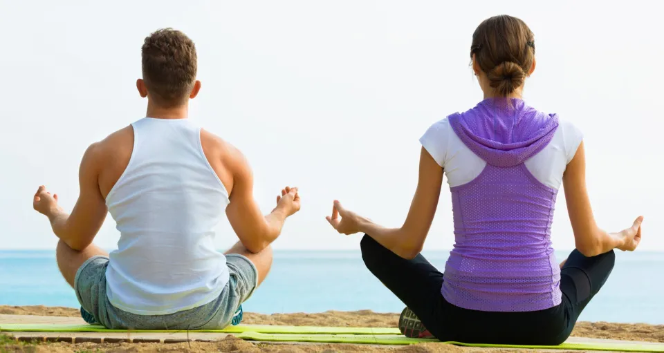 Glad woman and man sitting cross-legged do yoga poses on beach at daytime
